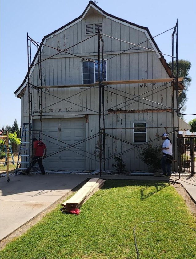 JJR Construction - Workers painting a two-story gray barn. Scaffolding surrounds the structure. The setting is a sunny yard.