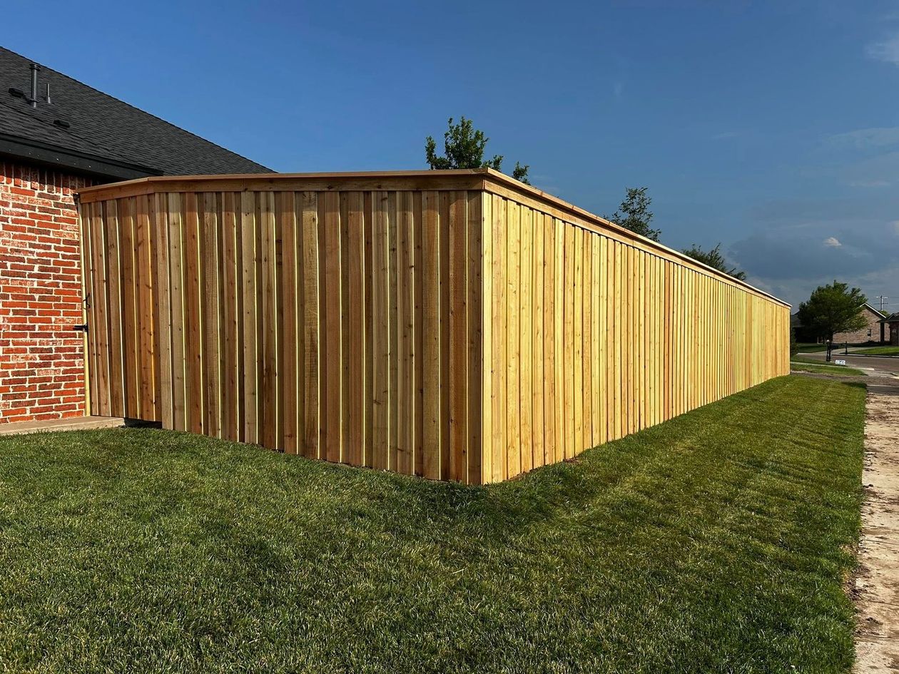 Wooden fence alongside a brick house and green lawn, under a blue sky.