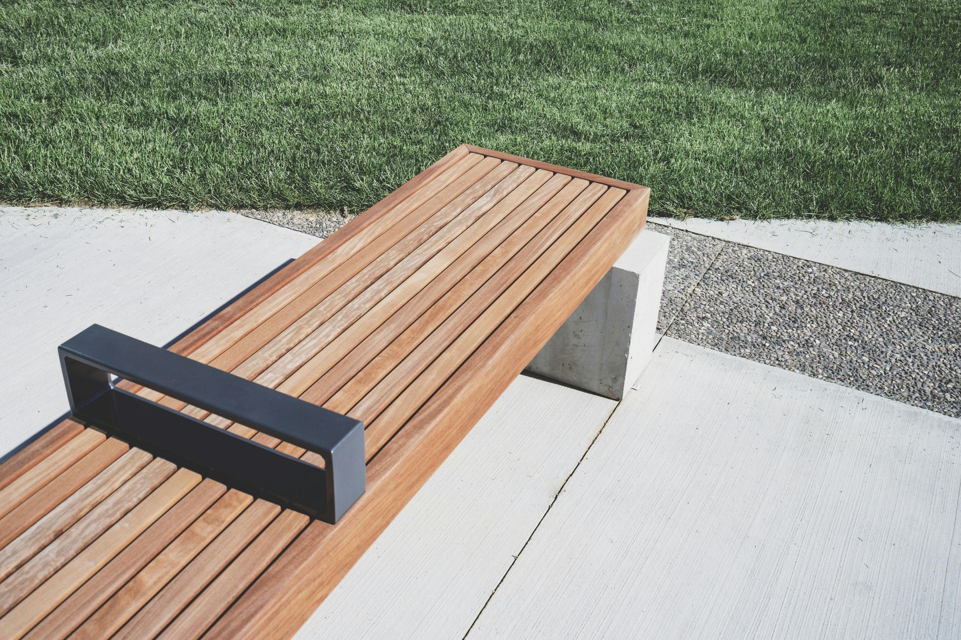 Wooden bench with a black armrest on a concrete slab, next to a patch of grass.