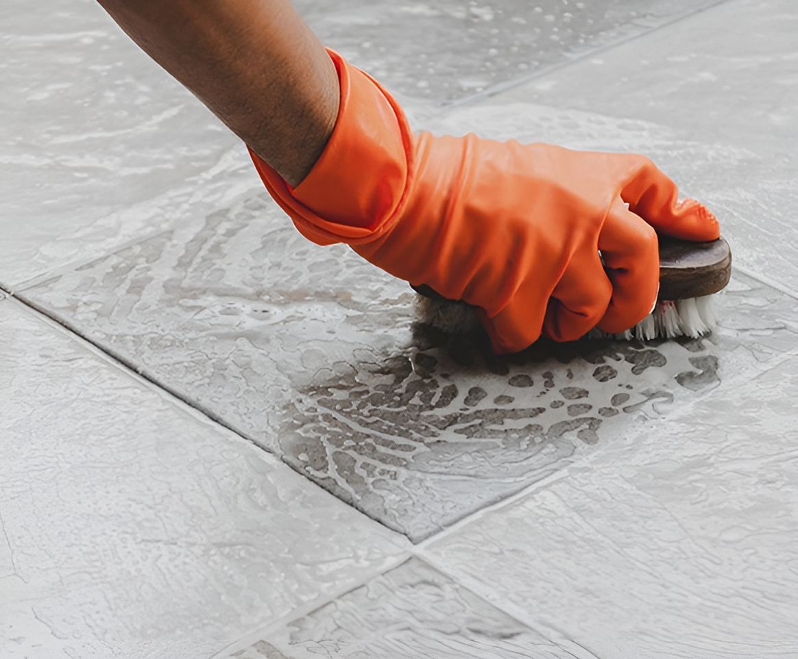 A Person Wearing Orange Gloves Is Cleaning A Tile Floor With A Brush — Regina's Cleaning Group Pty Ltd In Lee Point, NT