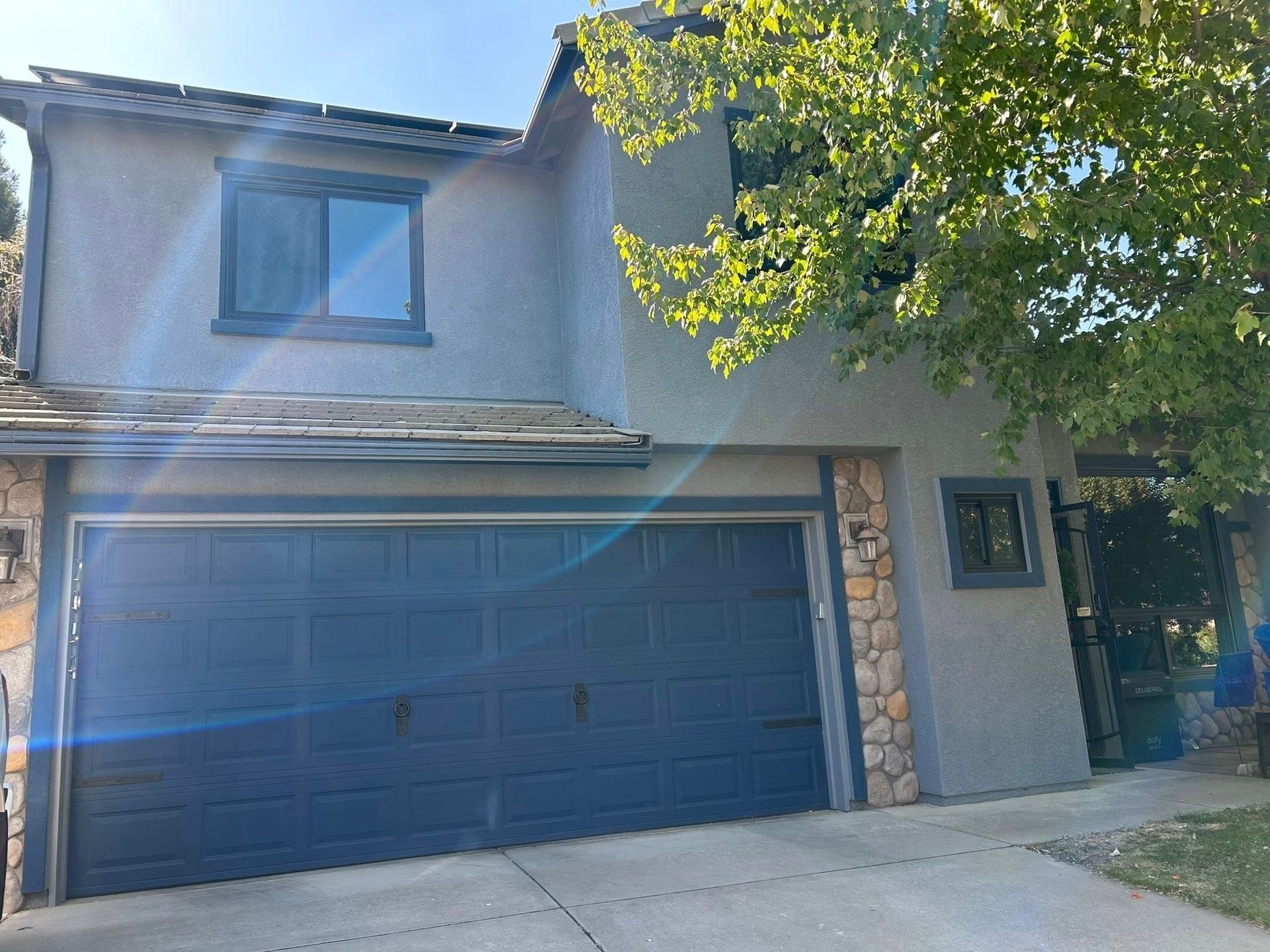 Two-story house with gray stucco exterior, blue garage door, and a tree on the right.