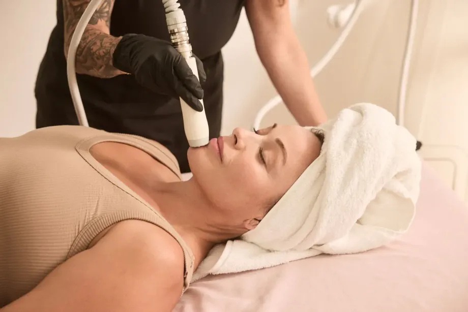 Woman receiving an Artemis T-Shape 2 facial treatment, lying on table; technician holding device. White towel on head.