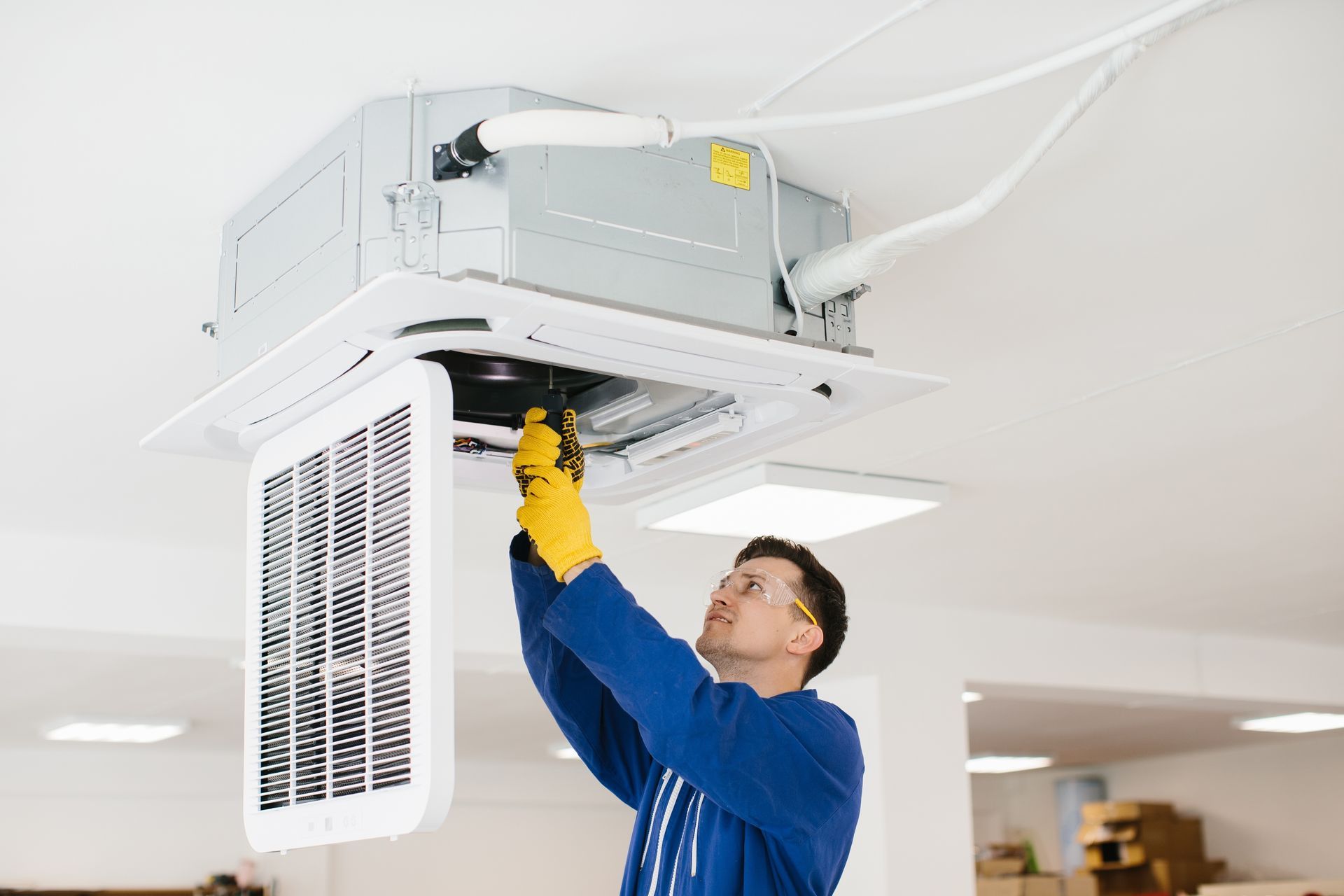 HVAC technician in blue jumpsuit, yellow gloves, and safety glasses, working on ceiling air conditioner unit.