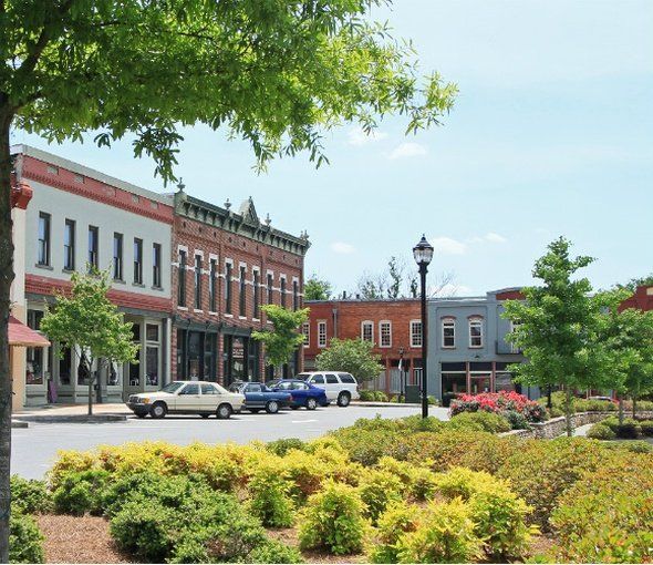 Street view of a small town with shops and cars parked along the street. Green trees and flower beds in the foreground.