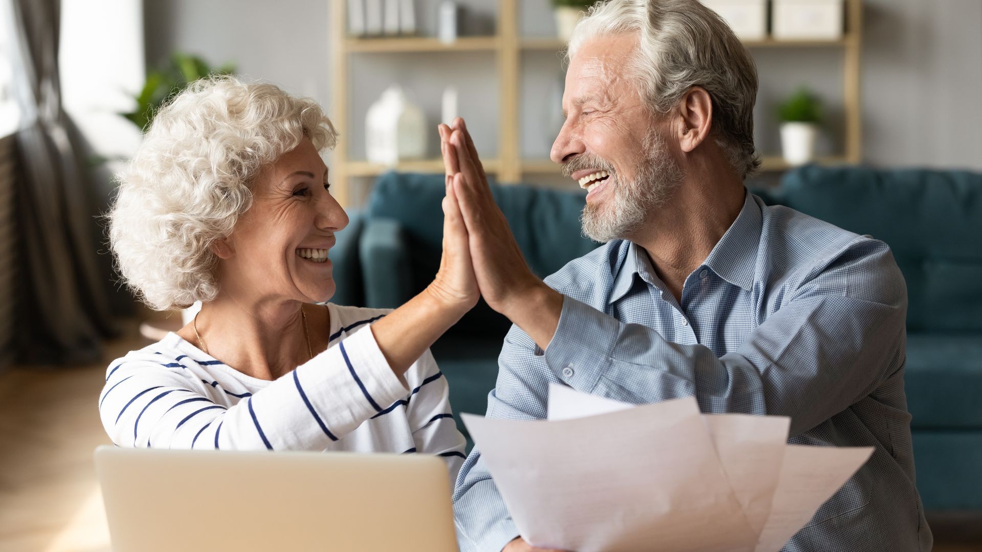 Senior couple high-fiving after reviewing paperwork in a living room, smiling.
