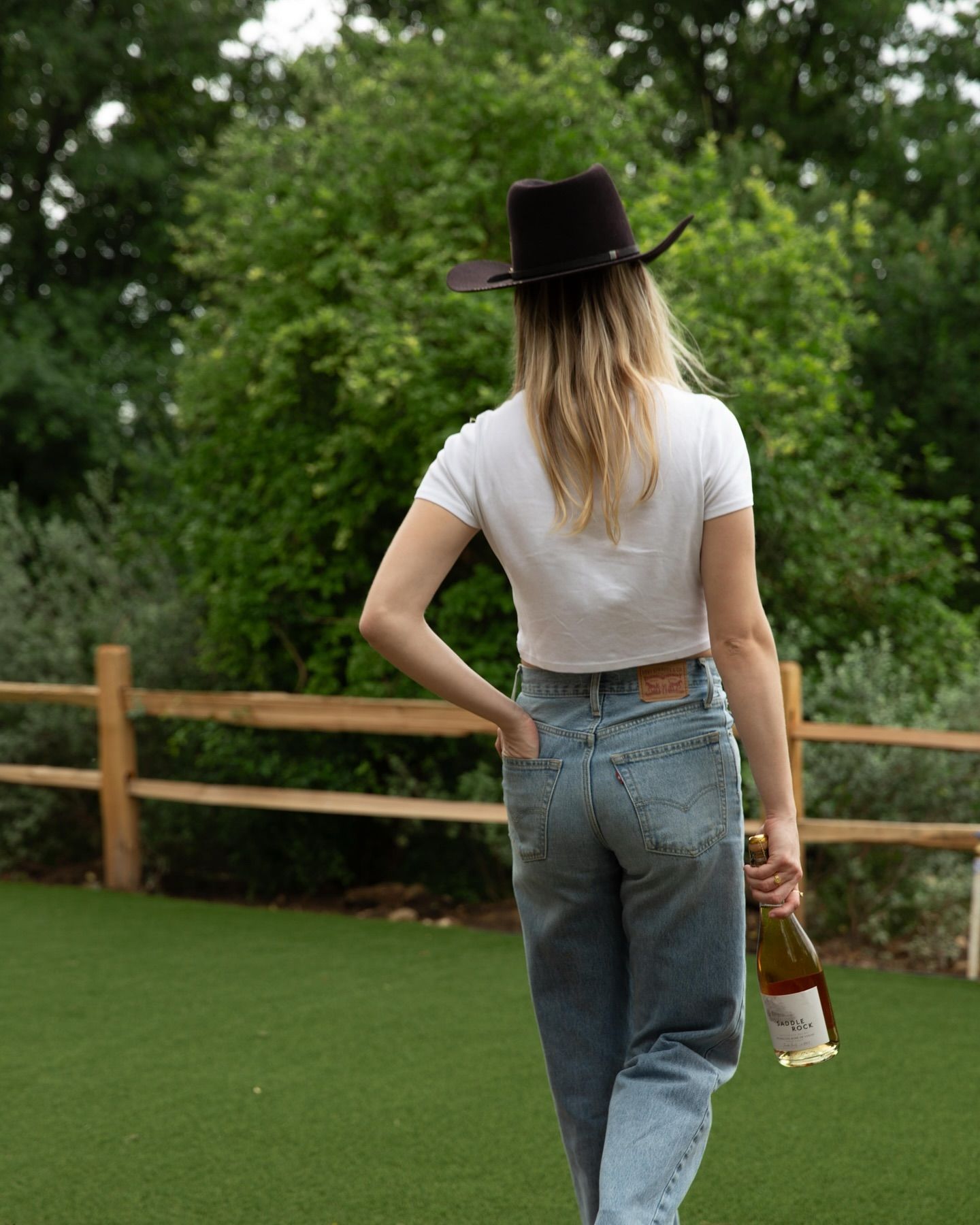 A woman wearing a hat and jeans is holding a bottle of wine.