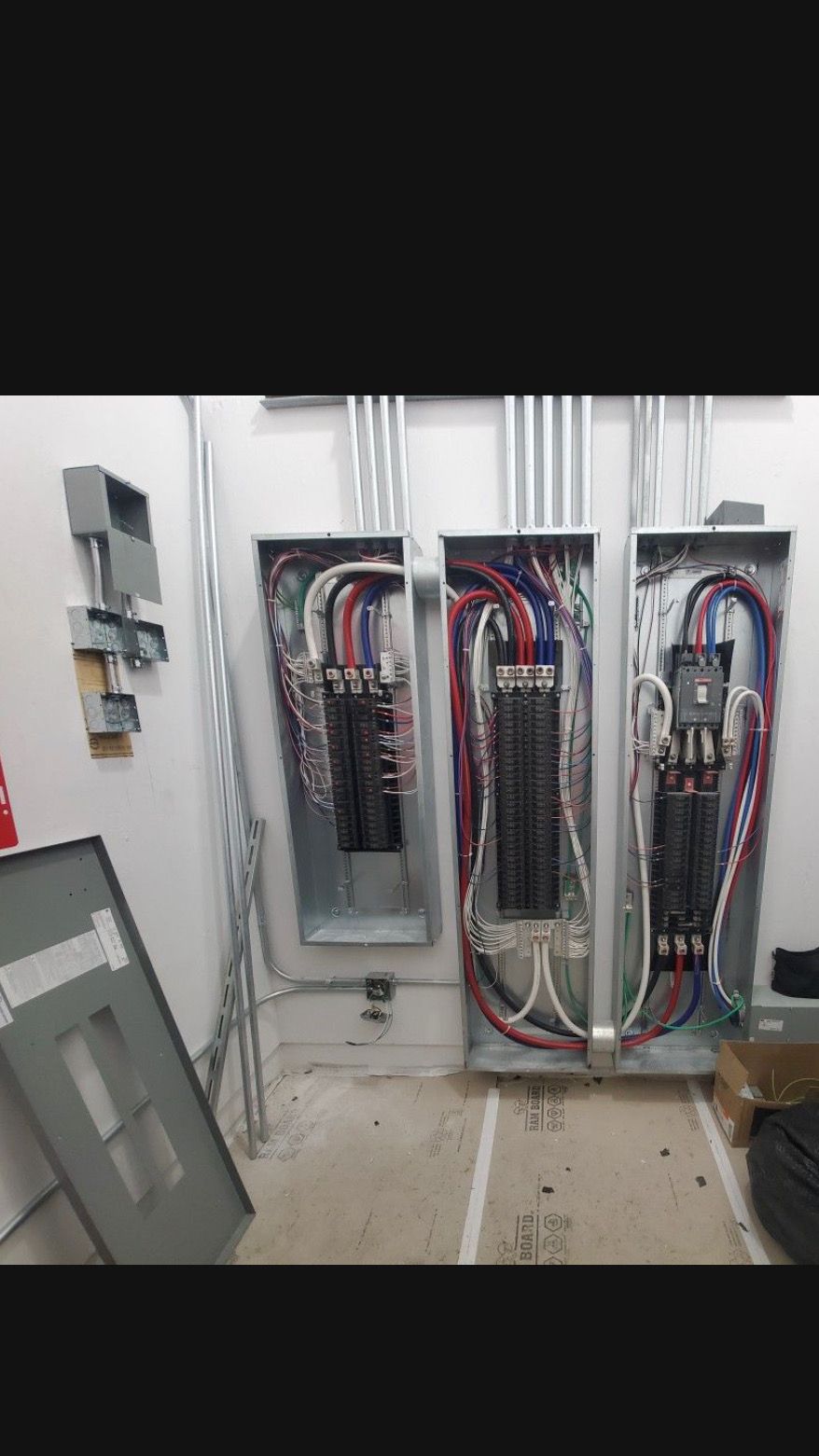 Three electrical panels installed on a wall with conduit and wiring visible.  A panel cover lies on the floor.