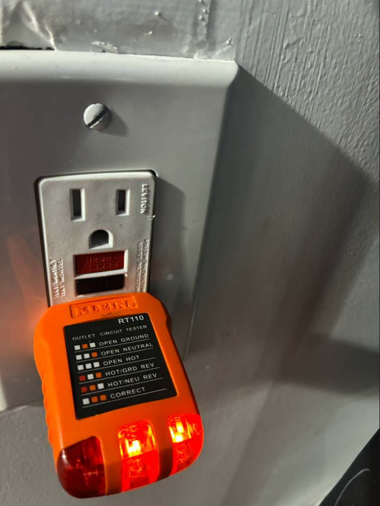 An orange electrical outlet tester plugged into a wall outlet, showing two red lights illuminated.