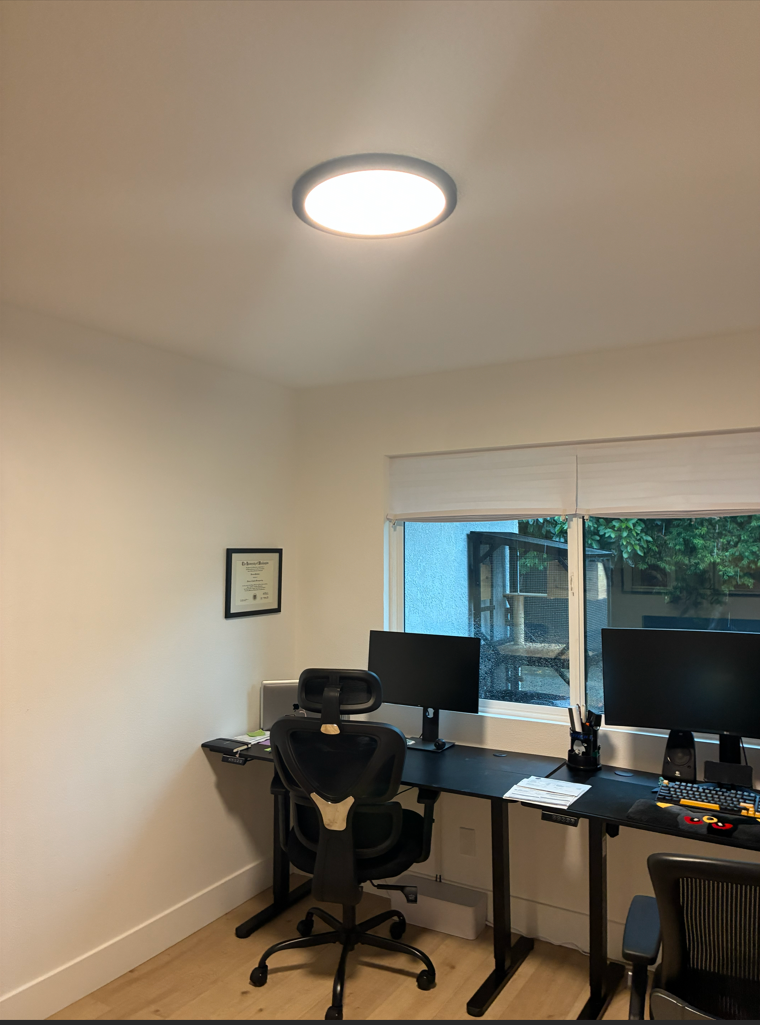 Office with a desk, two monitors, chair, and a bright overhead light. A window is in the background.