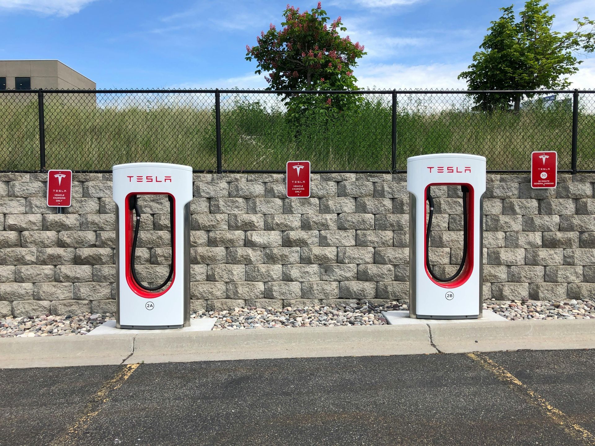 Tesla Supercharger stations against a brick wall, with charging cables and signs. Blue sky.