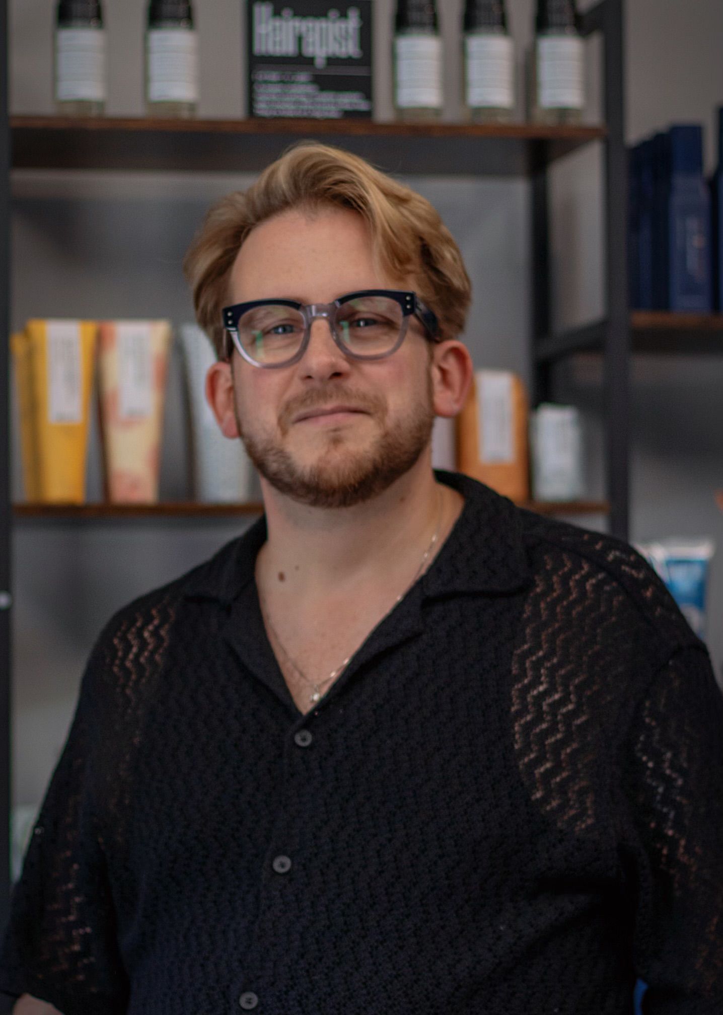 A man wearing glasses and a black shirt is standing in front of a shelf of hair products.