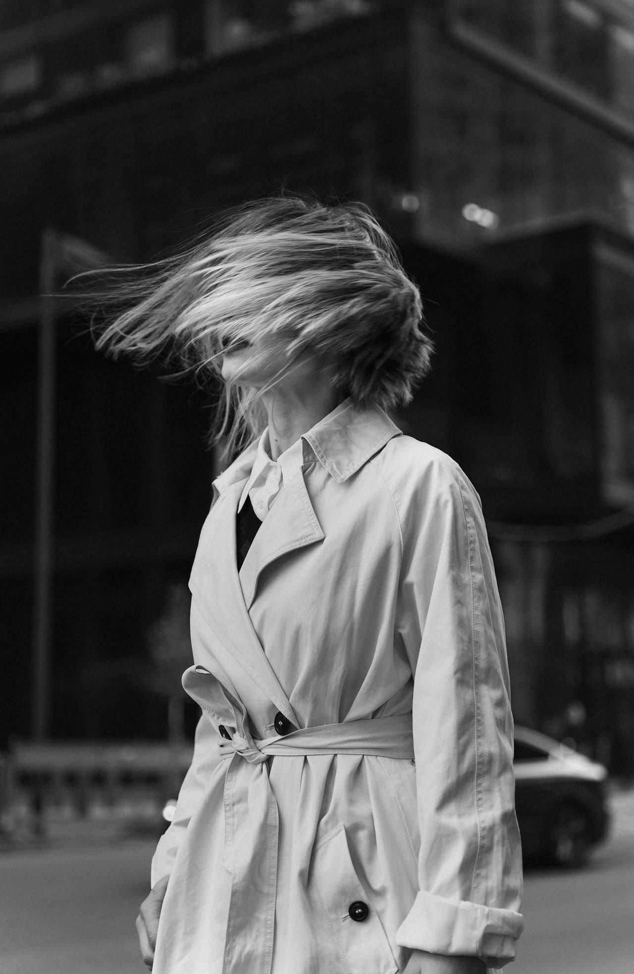 A black and white photo of a woman in a trench coat with her hair blowing in the wind.