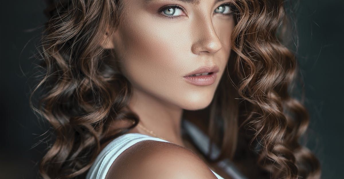 A woman with curly hair is wearing a white tank top and looking at the camera.