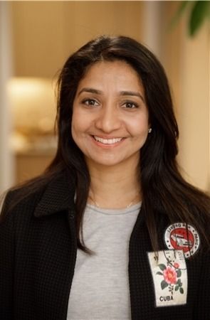 Woman with dark hair smiles, wearing a black jacket with patches in a room with light-colored walls.