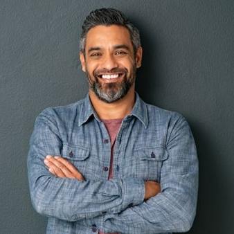 Man with graying hair and beard smiles, arms crossed, wearing blue button-down shirt, standing in front of gray wall.