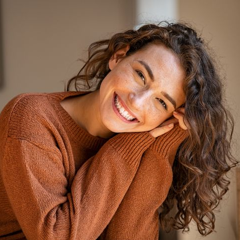 Woman in an orange sweater, smiling with head resting on arms. Curly brown hair, freckles, blurred background.