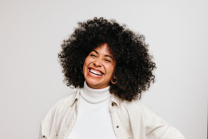 Woman with voluminous curly hair laughs, wearing a white turtleneck and light jacket against a white background.