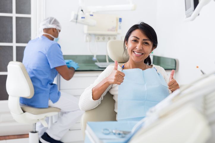 Woman in dentist chair gives thumbs up; dentist in scrubs prepares in background.