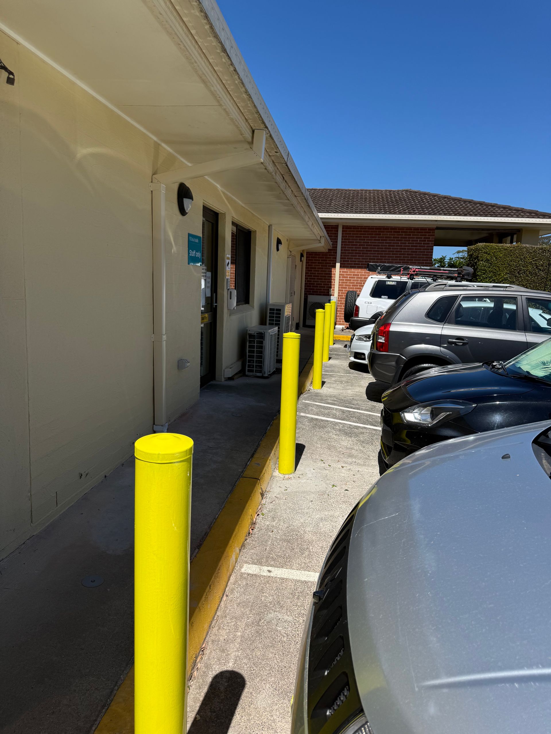 A row of bright yellow safety bollards lines the sidewalk next to a beige building and a parking area with several cars. — A1 Maintenance and Building In Old Bar, NSW