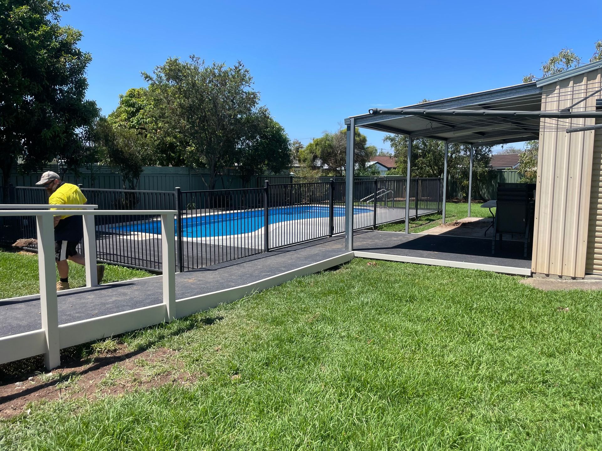 A person in a yellow shirt walks along a path next to a fenced outdoor swimming pool on a sunny day.A1 Maintenance and Building In Old Bar, NSW