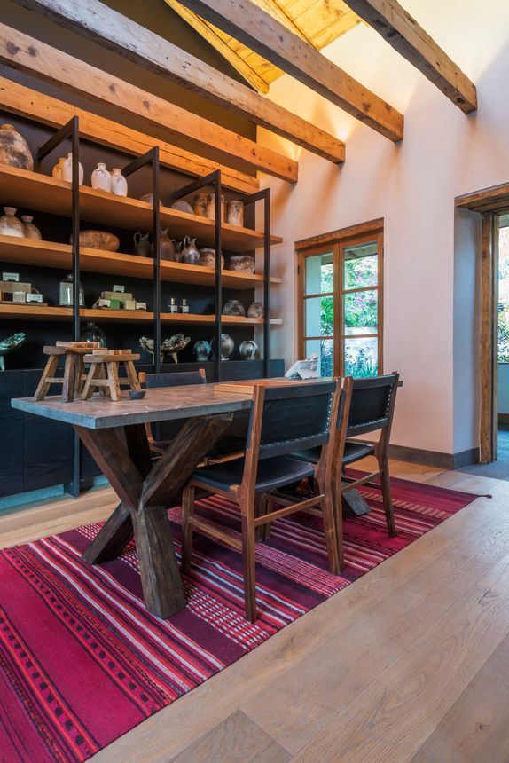 Comedor moderno con vigas de madera, una mesa larga, sillas, estantes y una alfombra roja estampada.