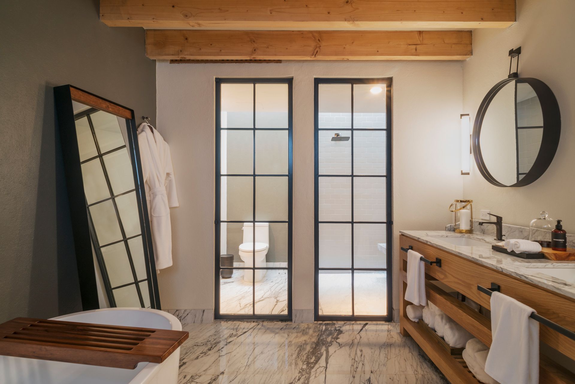 Baño estilo spa con puertas de ducha de cristal, tocador de madera, espejo redondo y vigas de techo de madera clara.