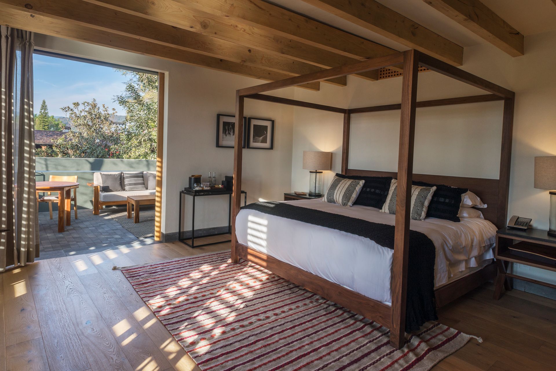 Dormitorio con cama con dosel, vigas de madera y puertas correderas que dan a un patio soleado.