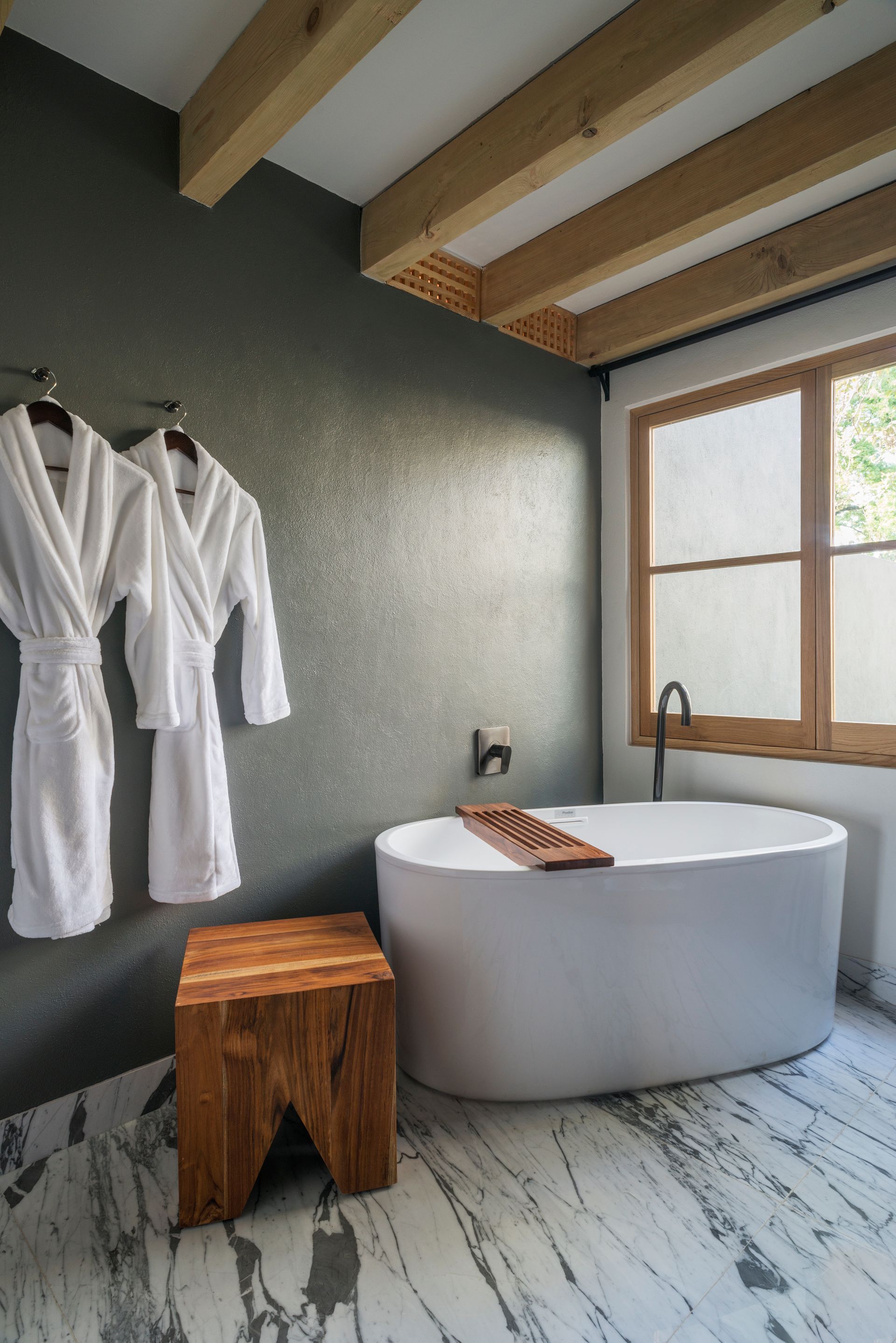 Baño minimalista con bañera exenta, taburete de madera, albornoces y una gran ventana bajo vigas vistas.