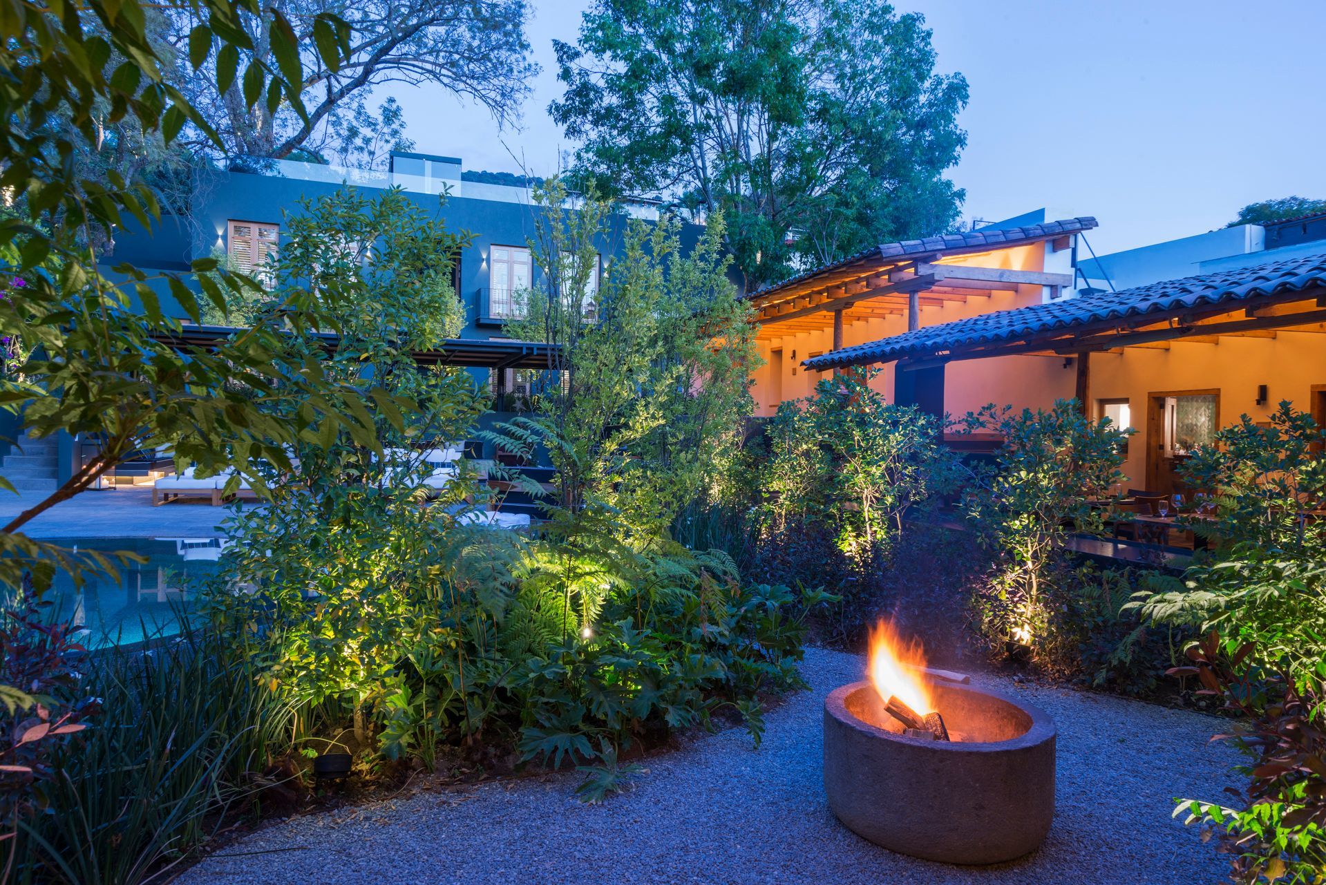 Patio ajardinado al atardecer con exuberante vegetación, luces cálidas y una hoguera en primer plano.