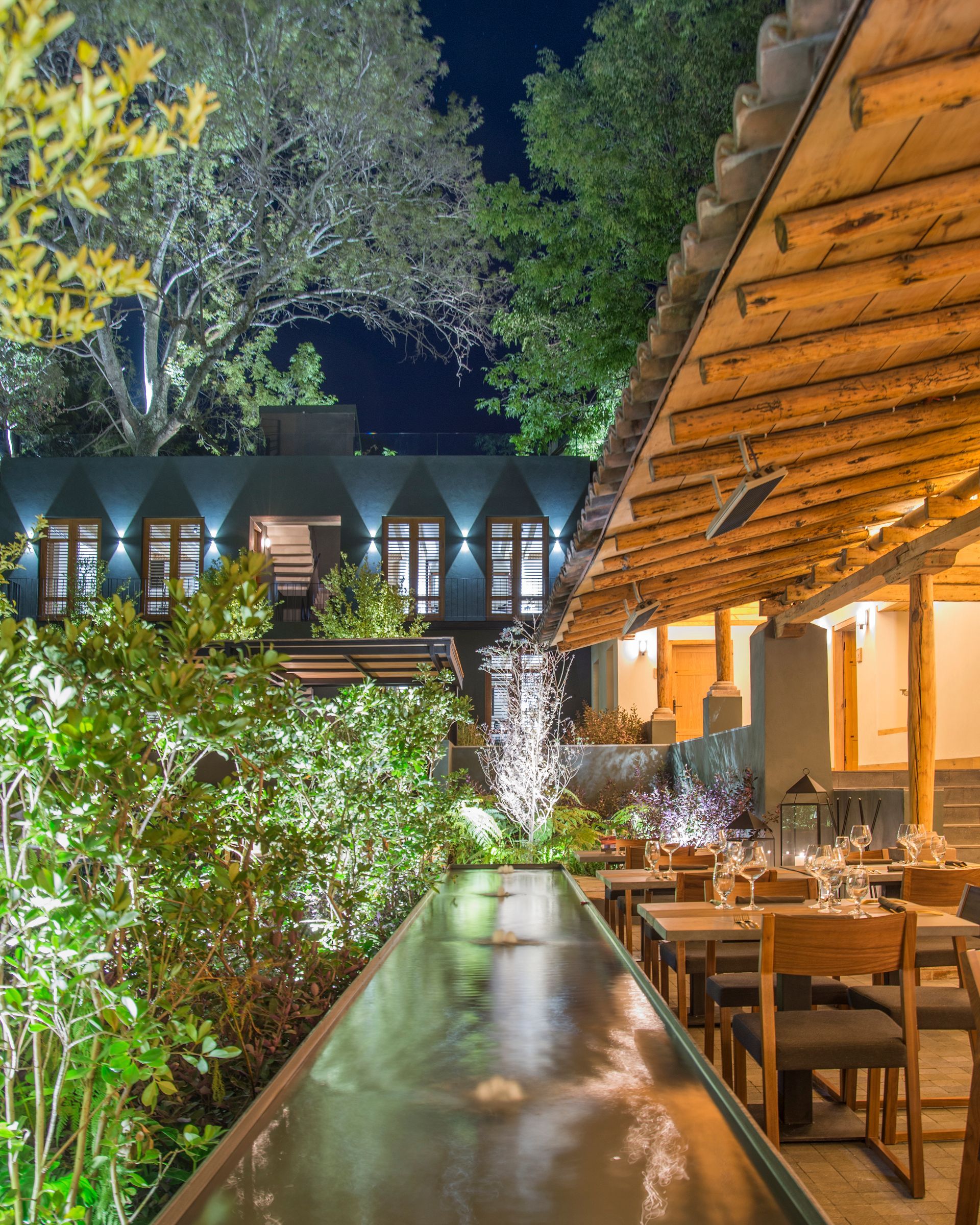 Terraza exterior de un restaurante con mesas de madera, vegetación y luces cálidas por la noche.
