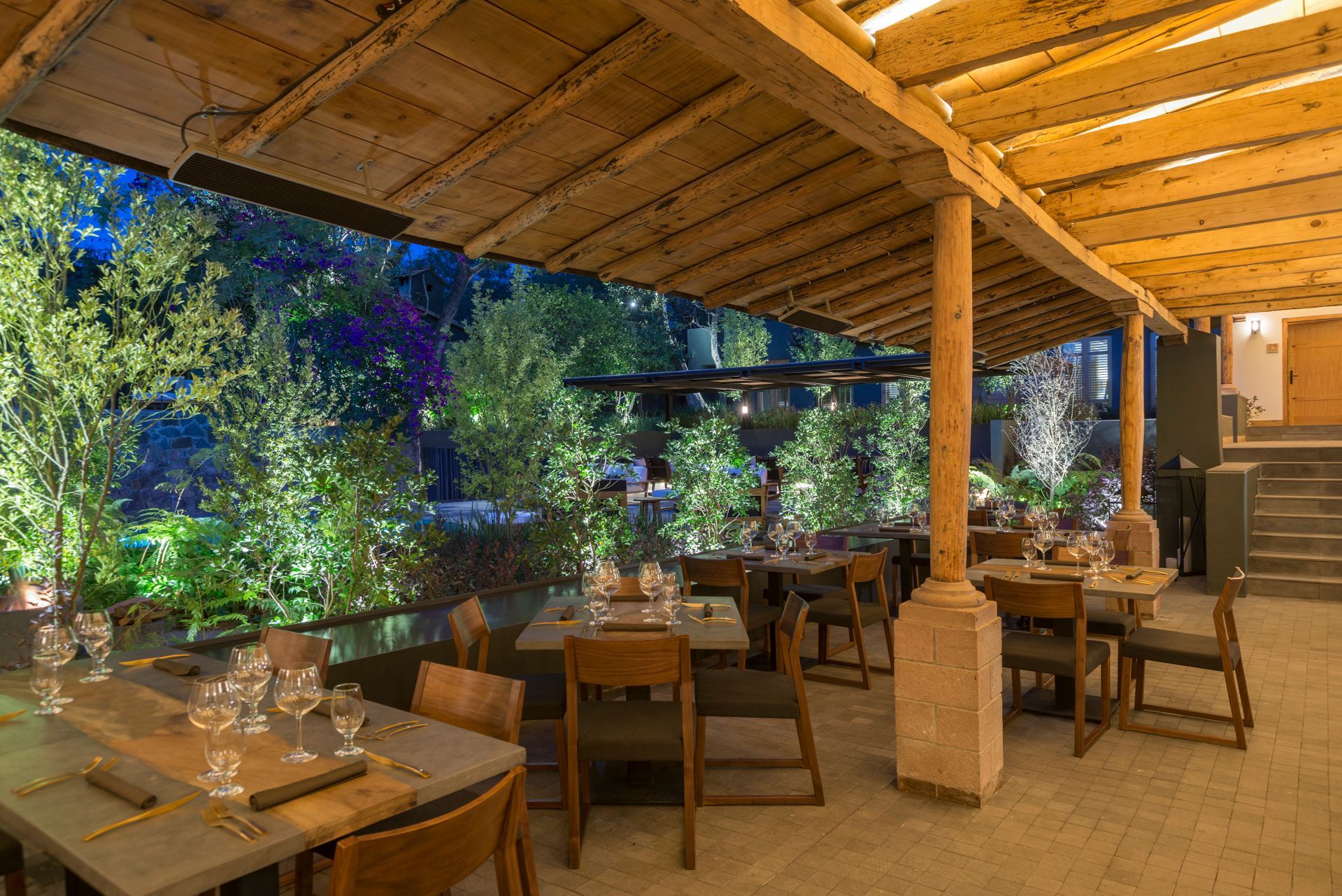 Acogedor patio exterior de restaurante con techo de madera, guirnaldas de luces, mesas y exuberante vegetación por la noche.