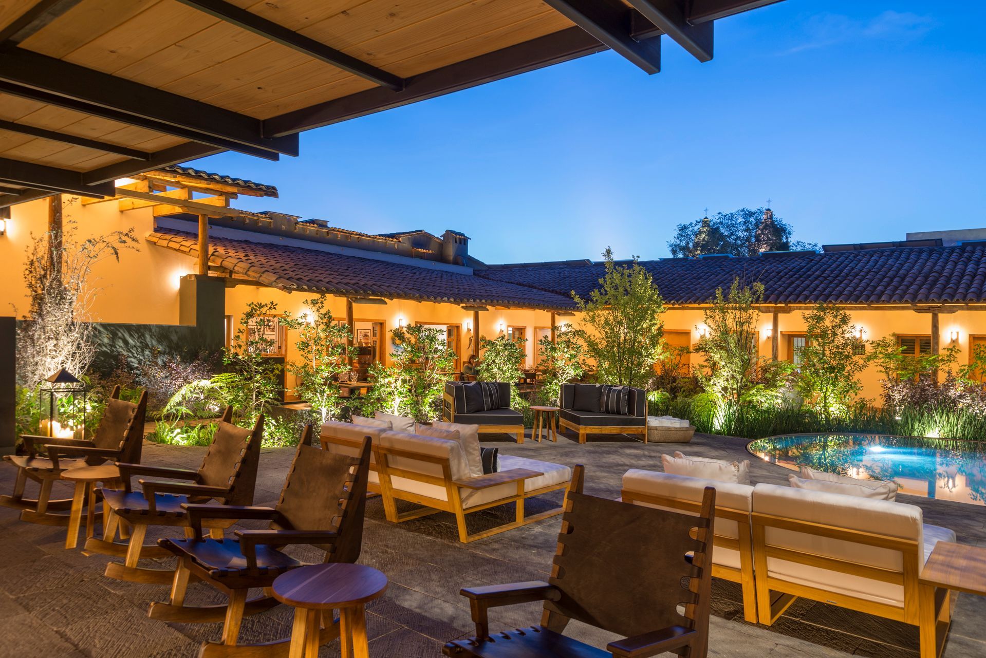 Terraza exterior de restaurante con mesas de madera, luces cálidas y exuberante vegetación al atardecer.