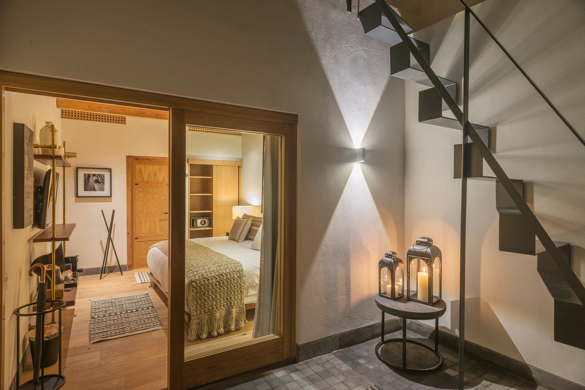 Dormitorio moderno visto a través de puertas correderas abiertas, con escaleras, faroles e iluminación ambiental cálida.