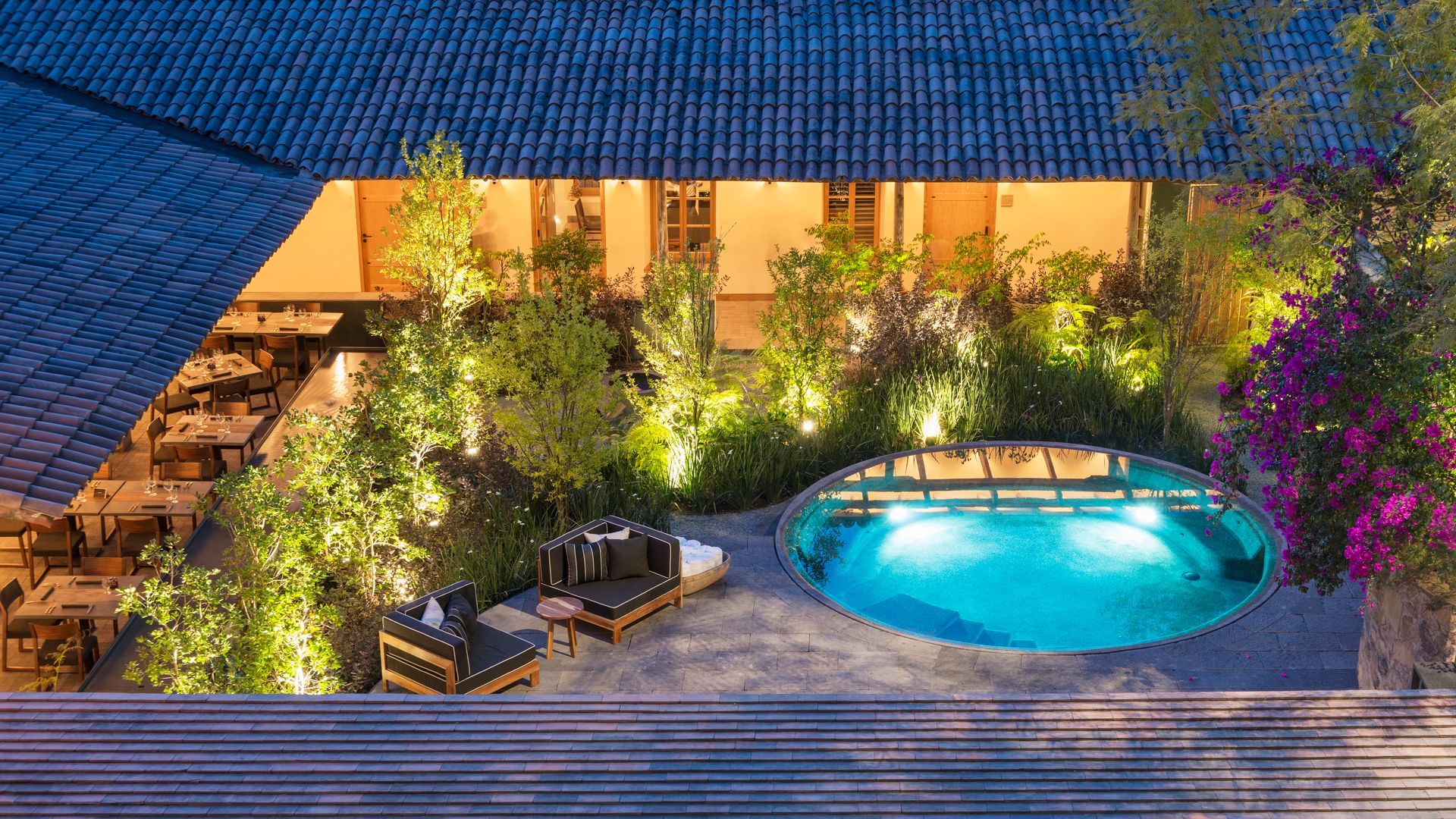 Patio junto a la piscina al atardecer, con una piscina turquesa iluminada, tumbonas y exuberante vegetación.