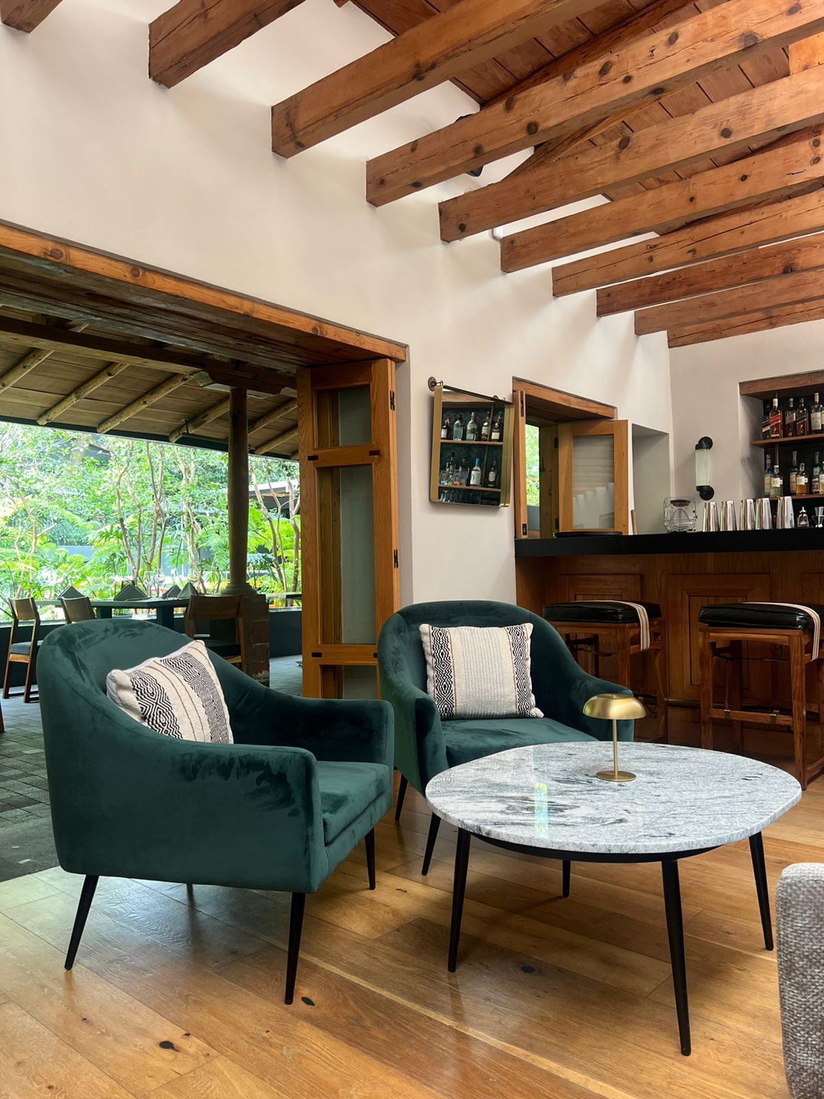 Acogedora sala de estar con sillones verdes, mesa de centro de mármol y techo con vigas de madera que da a un patio.