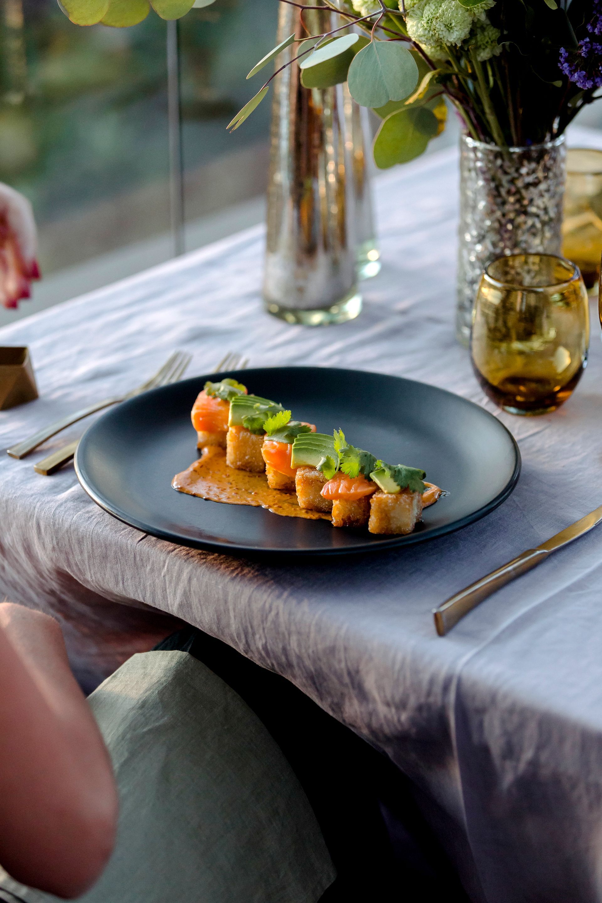Un aperitivo gourmet emplatado sobre una mesa con flores, copas y luz natural de fondo.