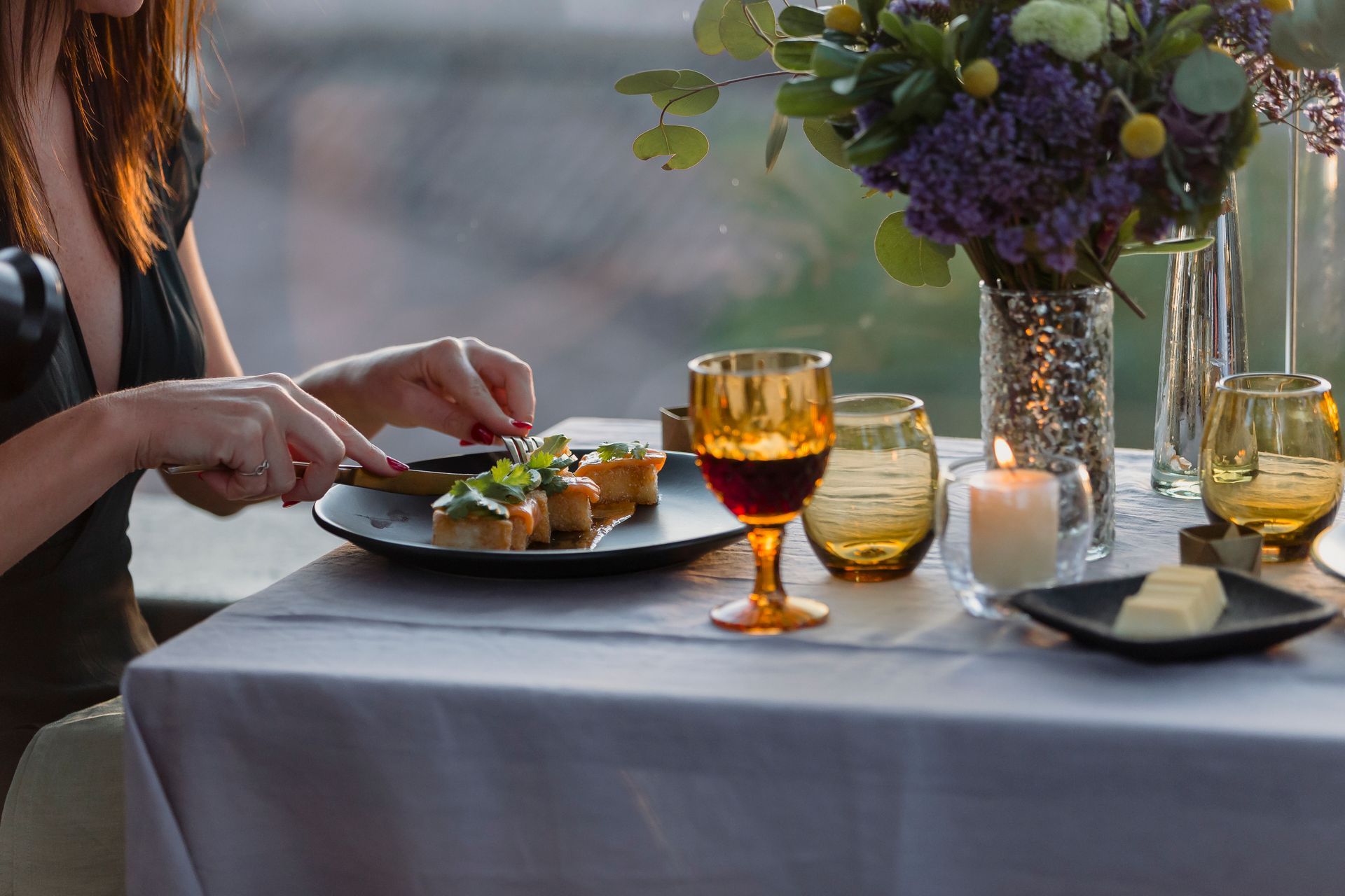 Una persona prepara la mesa para cenar al aire libre con copas de vino y un centro de mesa floral.