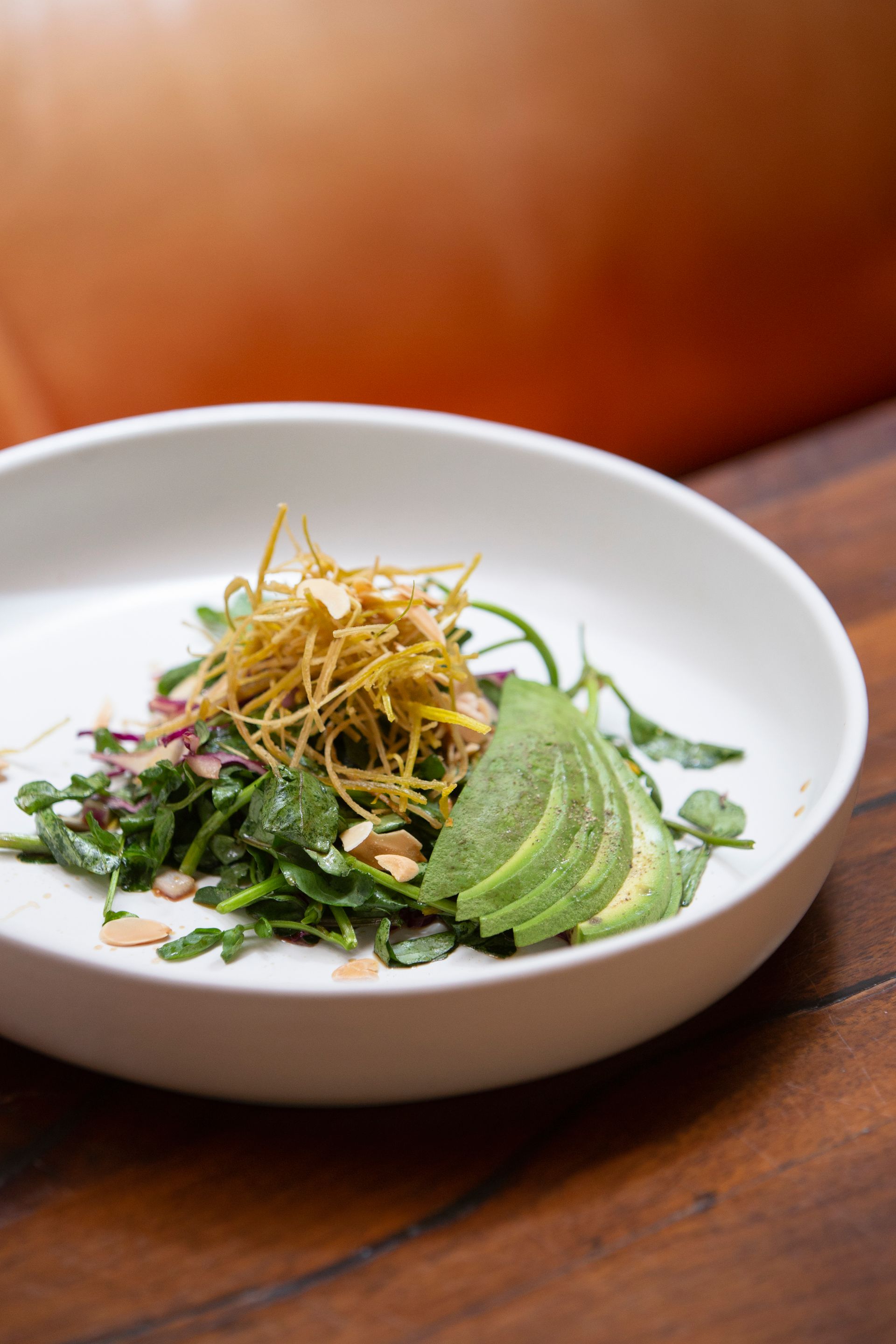 Bol blanco de ensalada verde con rodajas de aguacate y guarnición crujiente sobre una mesa de madera.