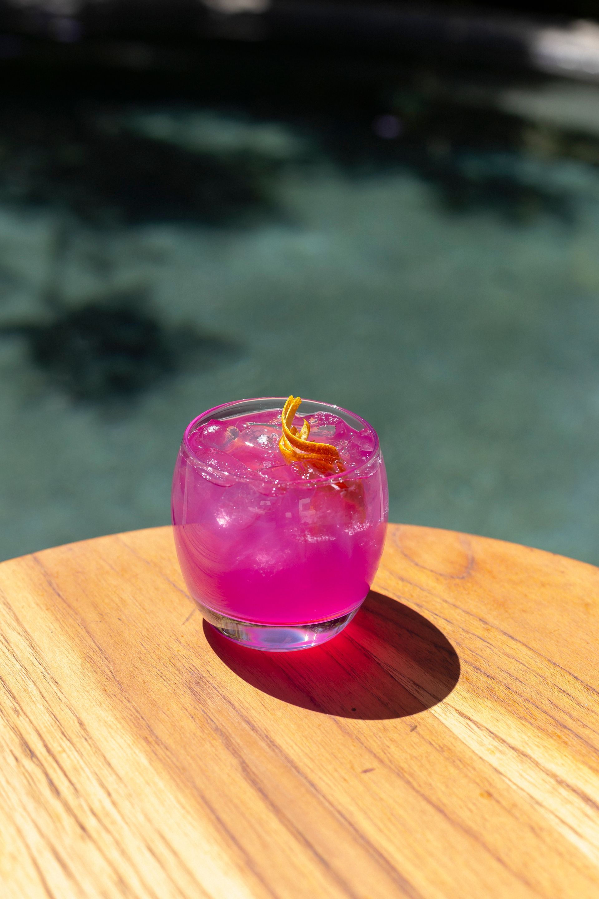 Bebida de color rosa brillante con guarnición de cítricos sobre una mesa de madera junto a una piscina.