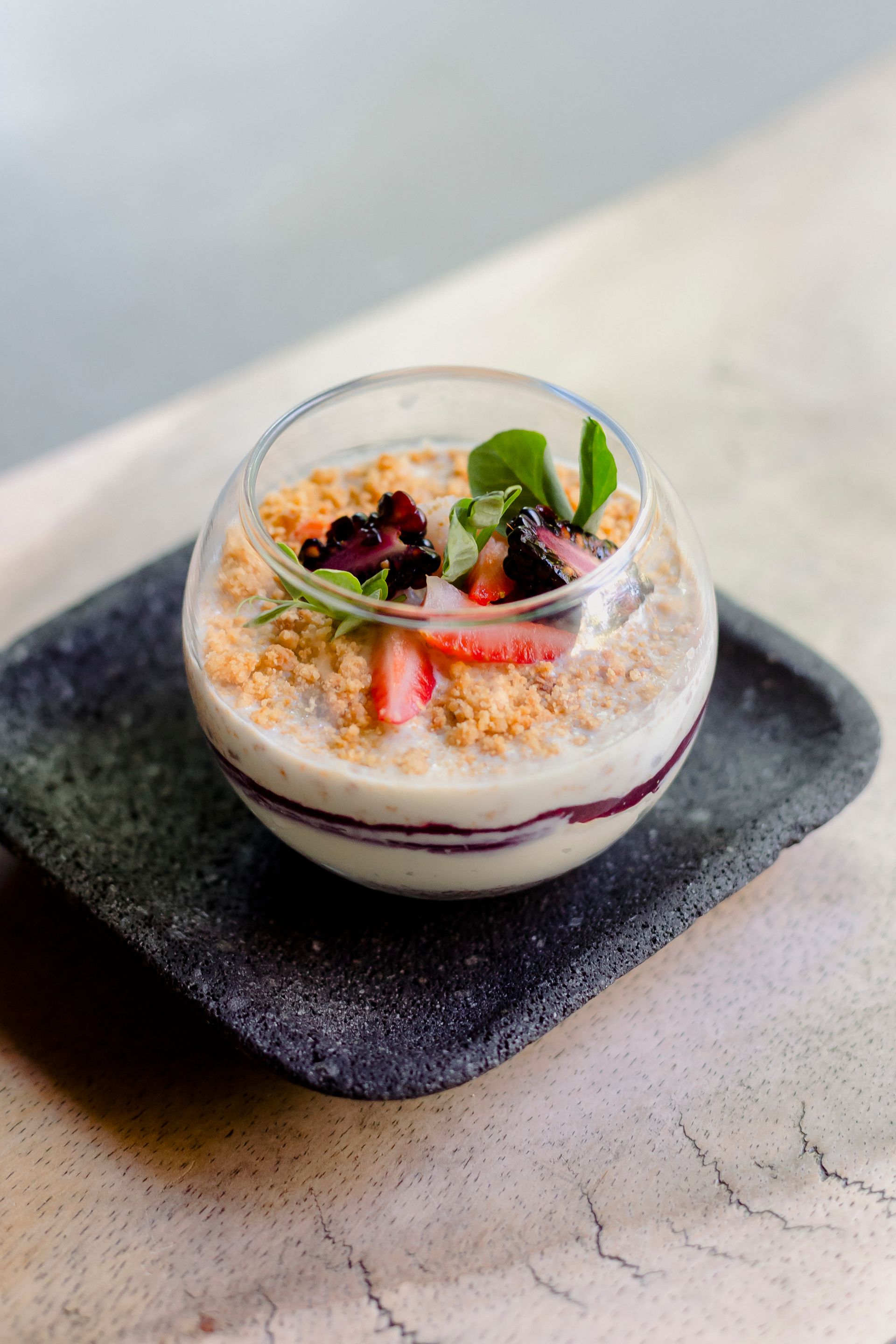 Vaso de postre de cristal con capas de yogur, granola, frutos rojos y menta sobre un plato de pizarra negra.