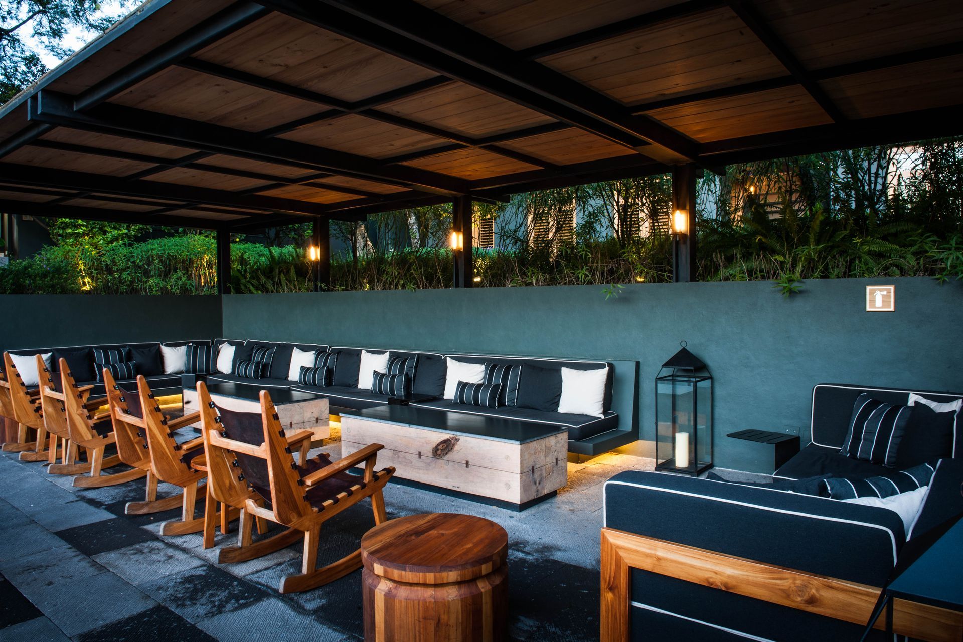 Patio al aire libre con sillas de madera, mesas bajas y bancos acolchados bajo un techo cubierto.