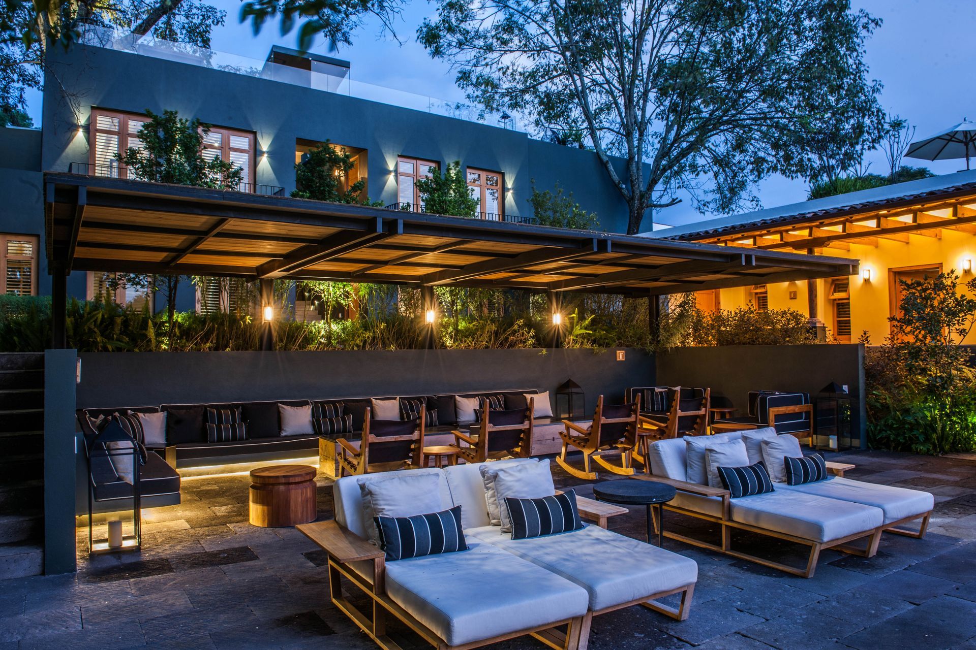 Salón al aire libre al atardecer con sofás acolchados, luces cálidas y una casa moderna al fondo.
