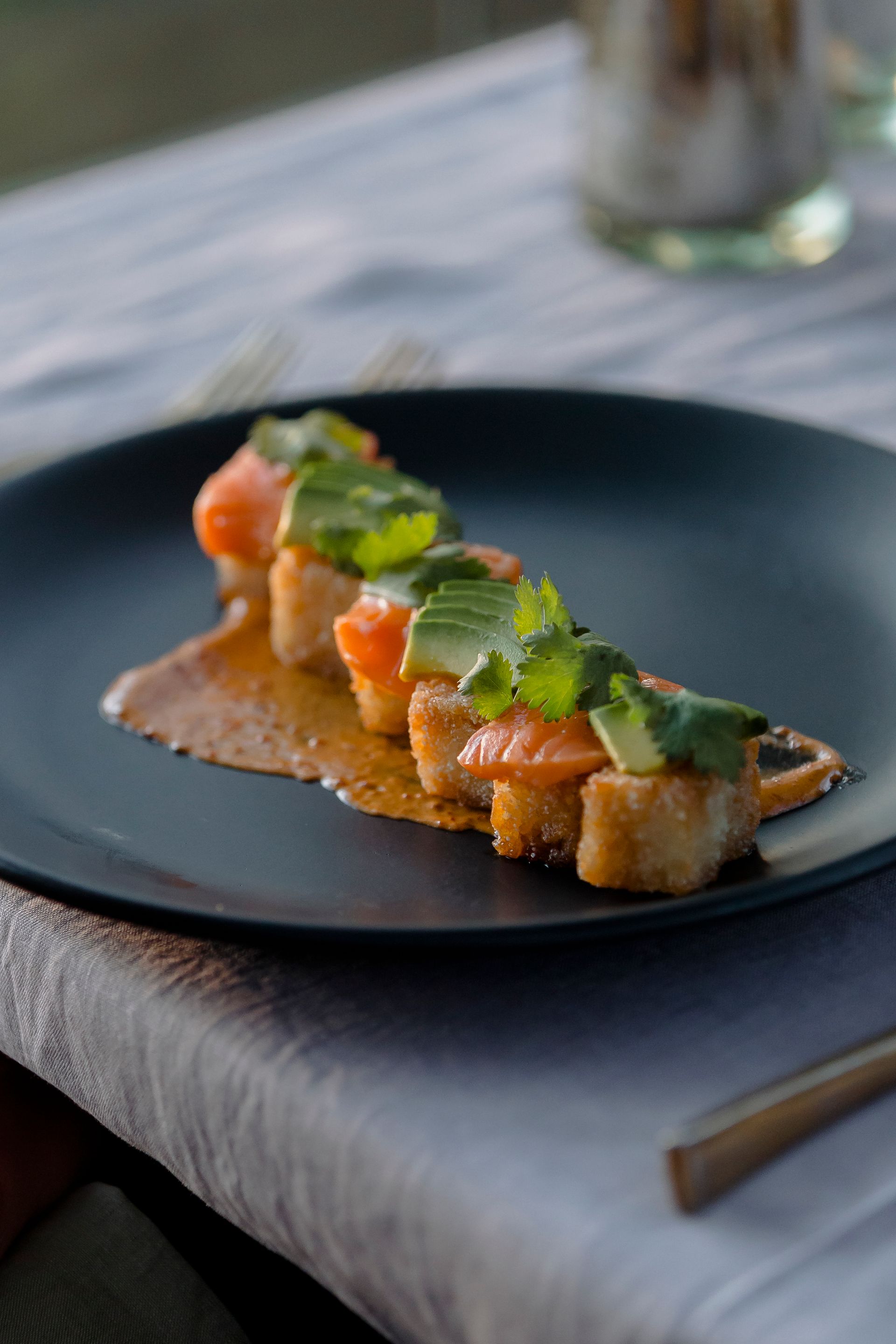 Elegante aperitivo de salmón emplatado en un plato negro con hierbas verdes y salsa de naranja sobre un mantel blanco.