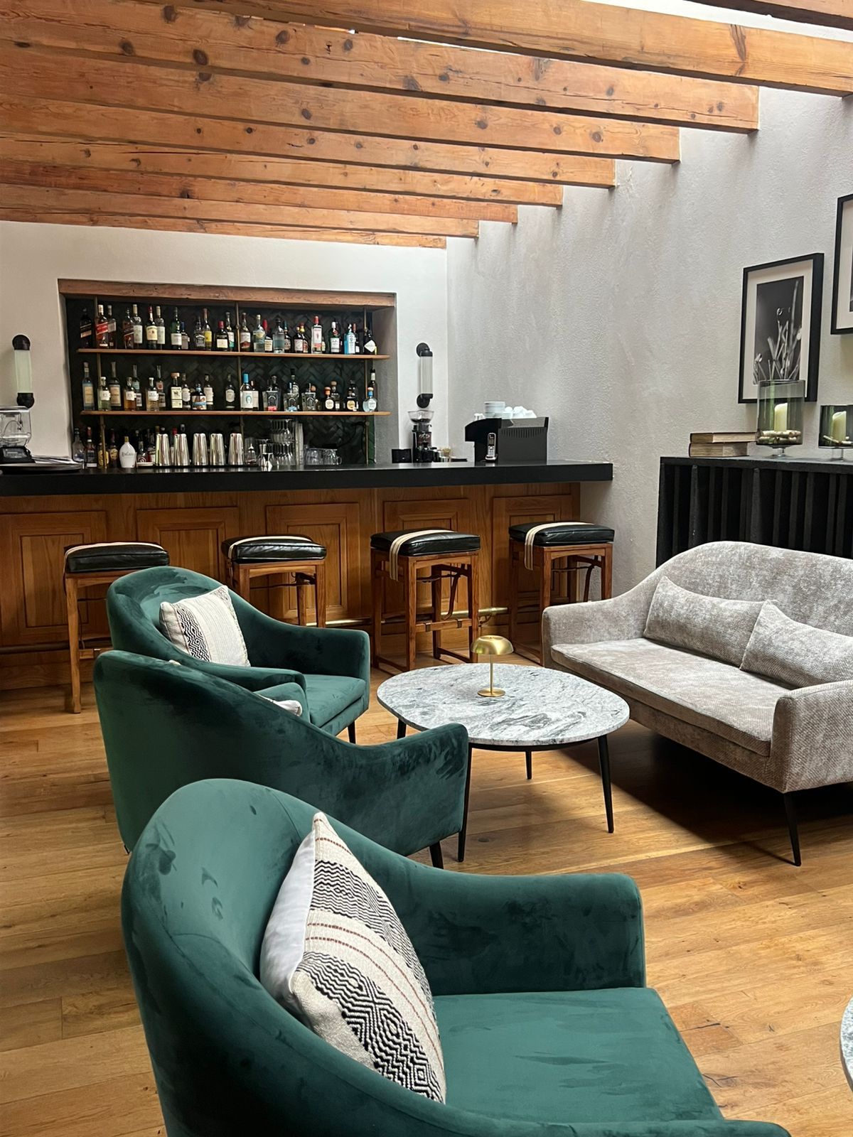 Acogedor salón con sillones de terciopelo verde, sofá beige, barra de bar y techo con vigas de madera.