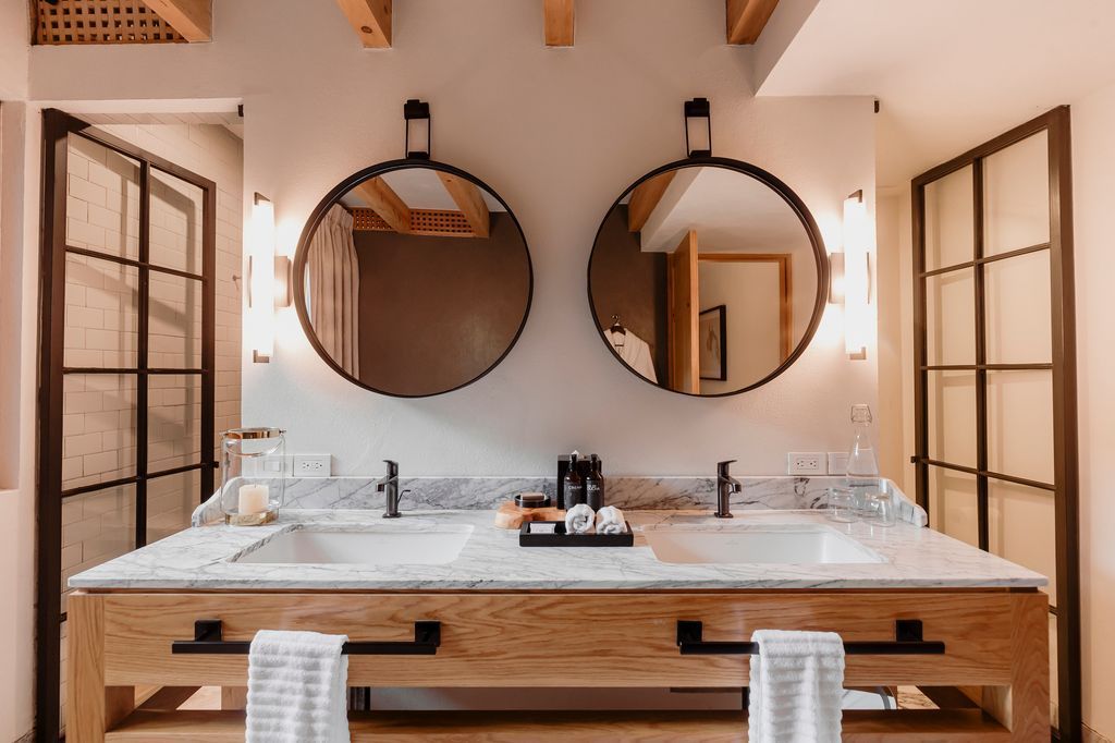 Tocador de baño moderno con dos espejos redondos, encimera de mármol y armarios de madera con iluminación cálida.