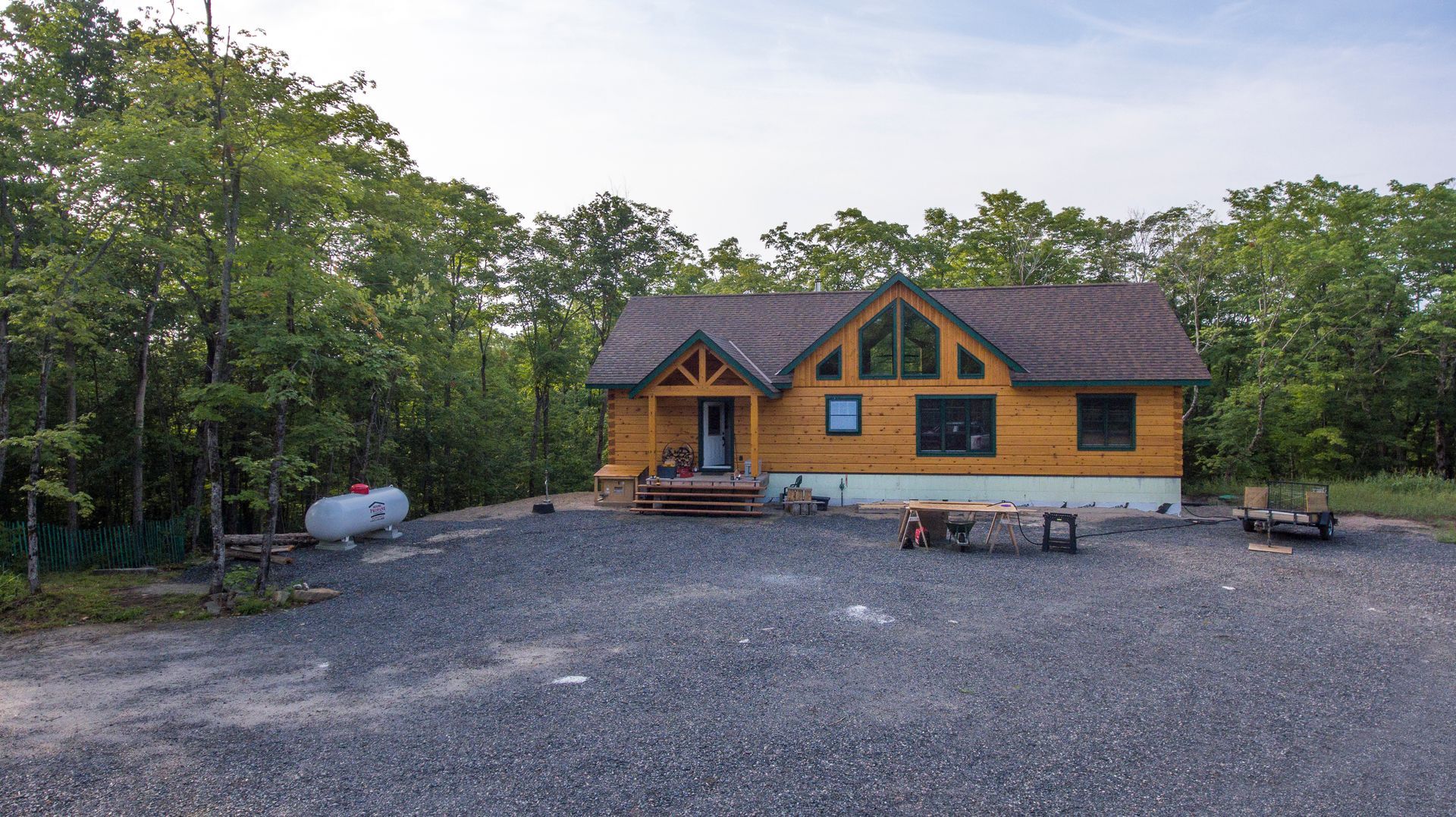 A log cabin in the middle of a forest with a gravel driveway in front of it.