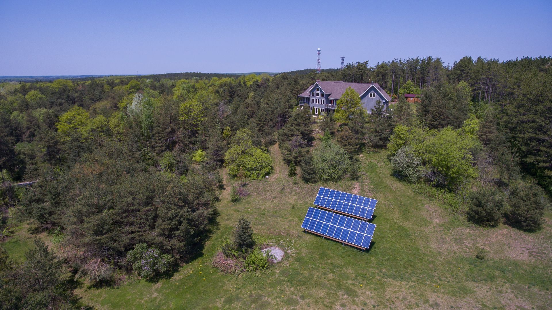 An aerial view of a house and a solar panel in a field surrounded by trees.