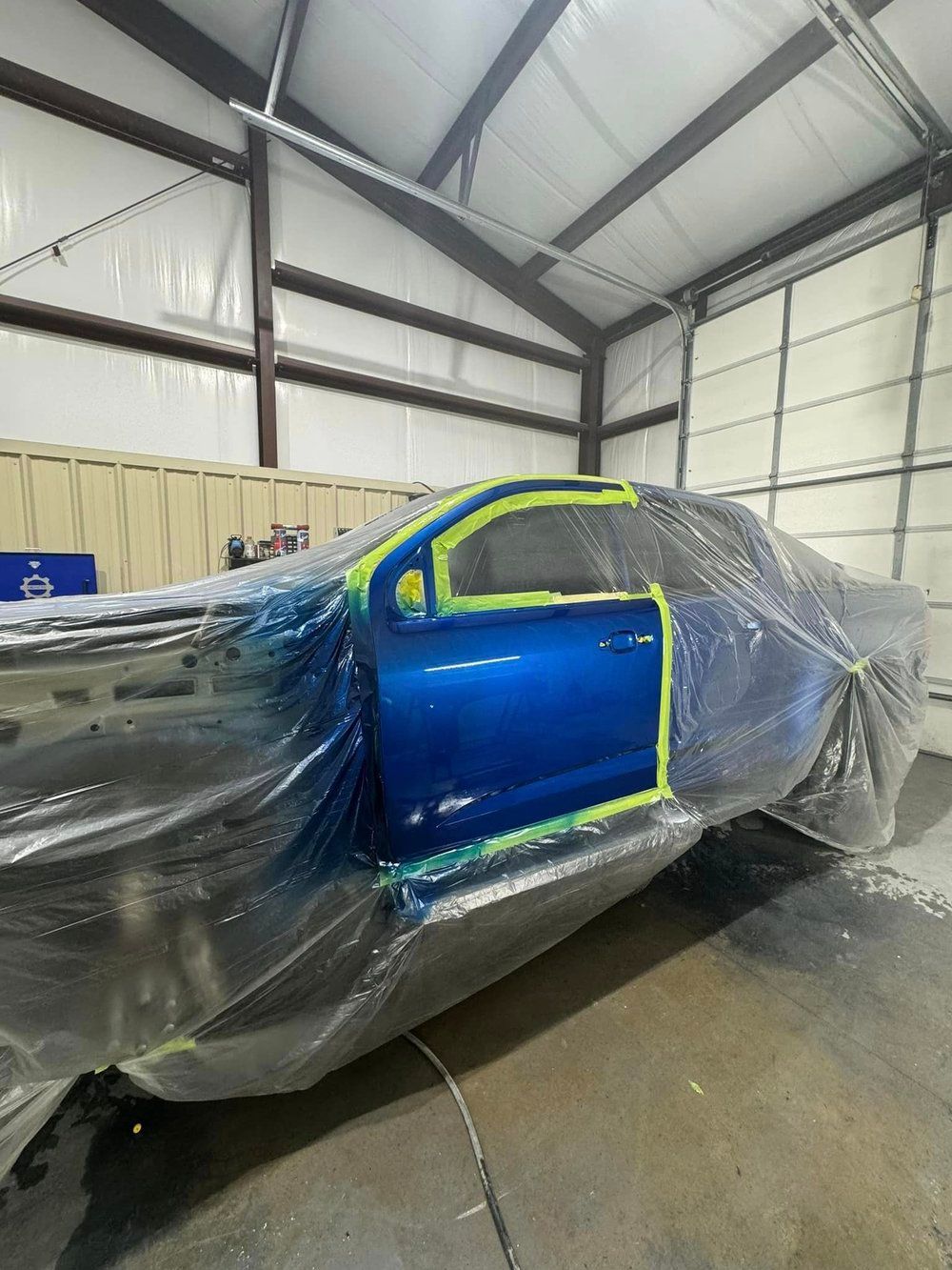 Blue vehicle door in a spray booth, taped and covered in plastic sheeting.