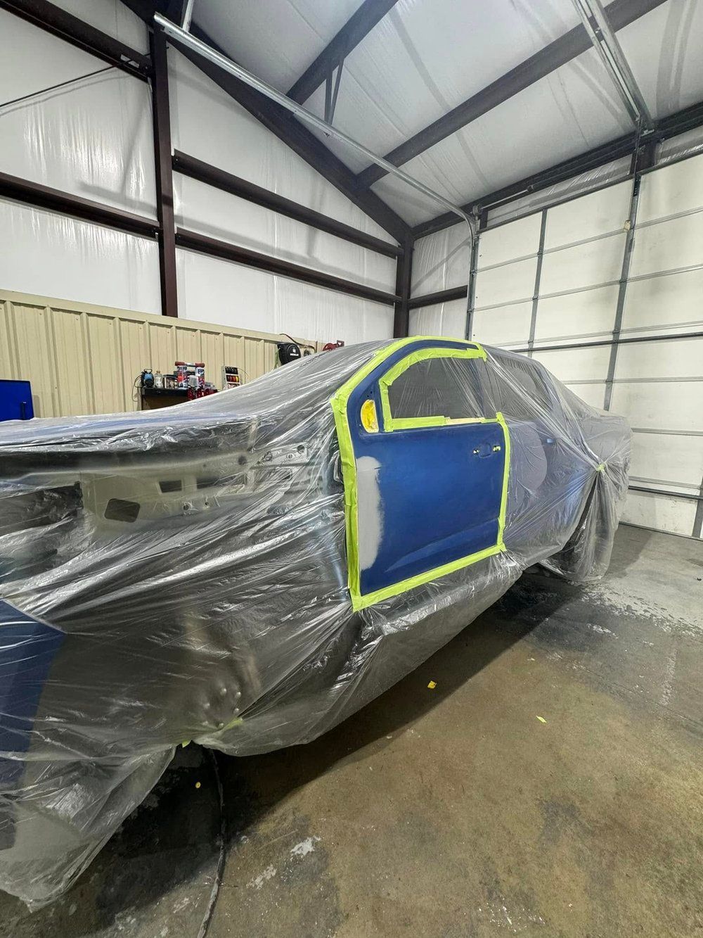 Car in a garage partially covered in plastic for paintwork; blue door with masking tape.