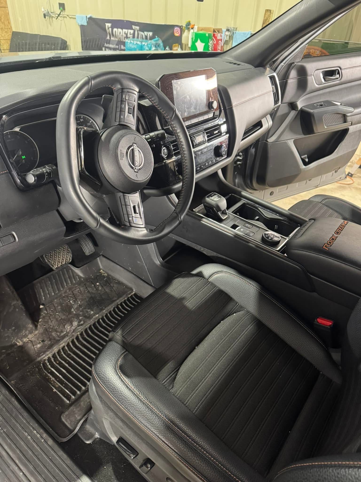 Interior view of a car with a black steering wheel, dashboard, and seat. The floor has black rubber mats.